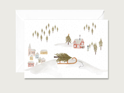 Handillustrierte Klappkarte mit einem Holzschlitten samt Tannenbaum vor verschneiten Dorfhäusern und Nadelbäumen neben weißem Umschlag auf hellem Untergrund.
