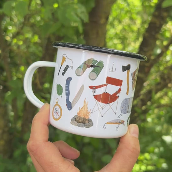 Eine Hand hält eine weiße Emaille-Tasse vor einem weich gezeichneten, grünen Waldhintergrund und dreht sie langsam um 360 Grad. Dabei wird das vollständige, umlaufende und detailreiche Design aus handgemalten Camping- und Naturmotiven wie Lagerfeuer, Rucksack, Bäumen und Outdoor-Ausrüstung sichtbar.