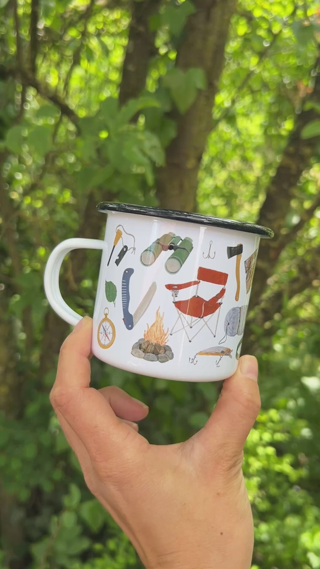 Eine Hand hält eine weiße Emaille-Tasse vor einem weich gezeichneten, grünen Waldhintergrund und dreht sie langsam um 360 Grad. Dabei wird das vollständige, umlaufende und detailreiche Design aus handgemalten Camping- und Naturmotiven wie Lagerfeuer, Rucksack, Bäumen und Outdoor-Ausrüstung sichtbar.